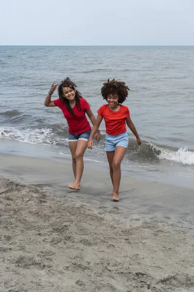 Okyanus kıyısında yürüyen afro saçlı iki gülümseyen kız.