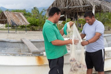 İki balıkçı birlikte çalışarak tropik bir sahilde bir teknenin yanında balık ağı hazırlıyor.