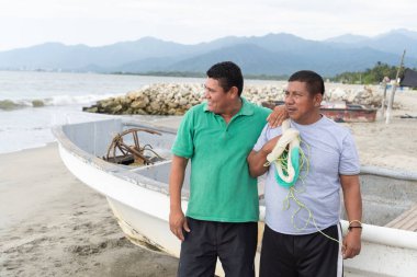 İki Latin Amerikalı balıkçı bir kumsalda yan yana durur, dostluk ve balık ağı gösterirler.