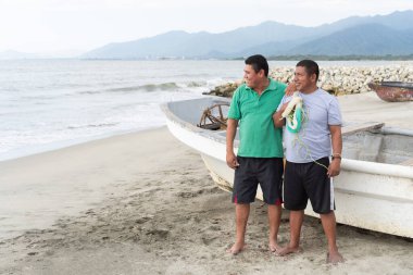 İki balıkçı, kumlu bir sahilde bir teknenin yanında elinde balık ağıyla, gülümsüyor.