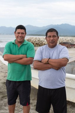 Sahilde duran iki balıkçı. Arka planda bir tekne ve dağlar var.