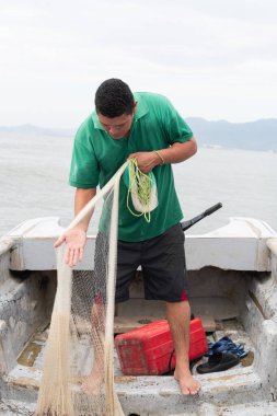 Balıkçı teknede çıplak ayakla, elinde balık ağıyla, çalışmaya hazırlanıyor.