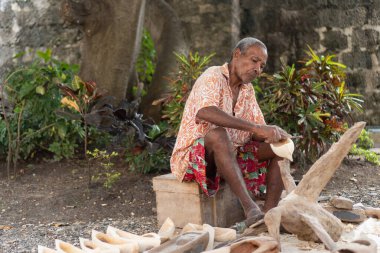 Yaşlı bir adam ahşap bir nesneyi açık havada keskiyle şekillendiriyor.