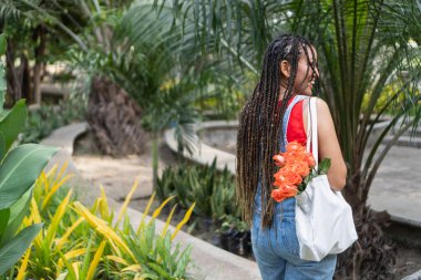Saç örgülü bir kadın çantasının içinde turuncu güller taşıyor, tropik bir bahçede mutlu mesut yürüyor.