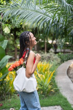Saçları örgülü bir kadın çantasında çiçeklerle bir parkta yürürken mutlu bir şekilde gülümsüyor.