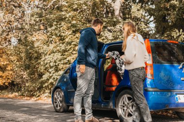 Erkek ve kadın, köpekleriyle dışarıda, arabanın yanında eğleniyorlar.