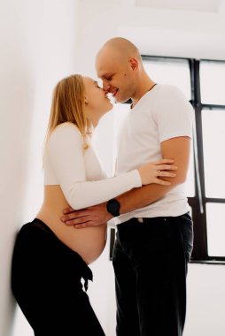 Mutlu genç çift, evde pencerenin önünde bebek beklerken sevgi dolu koca, hamile karısının karnına şefkatle dokunuyor..