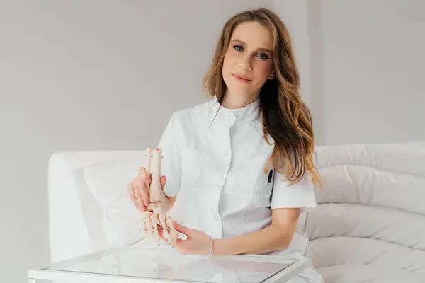 A doctor studies a womans leg structure while holding model of skeleton on a white table indoors. Models of skeletons help doctors study the structure of the human body.