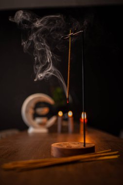 Smoke curls of incense stick in wooden holder for relaxation and meditation exercises, black background. Aromatherapy session with burning aroma stick, pleasant aroma and incense.