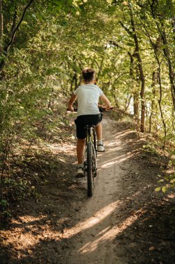 Genç bir çocuk güneşli, çamurlu bir patika boyunca bisikletini sürüyor yemyeşil bir ormanda kıvrılarak.