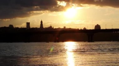Akşam şehrinin silueti karanlığa gömülüyor. Ukrayna 'nın başkenti Kyiv panoramik bir köprü manzarası, Dnipro nehrinin suyu, sarı günbatımına karşı binalar. Bir metropolde ışıklar sönüyor, elektrik kesiliyor.