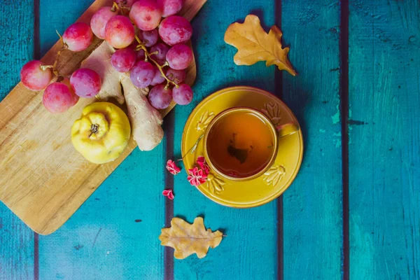 Beautiful fall still life with yellow cup on saucer. Bunch of red grapes, ripe agave, ginger root lie on cutting board. Fallen oak, maple leaves. Wooden green emerald background top view flatly.
