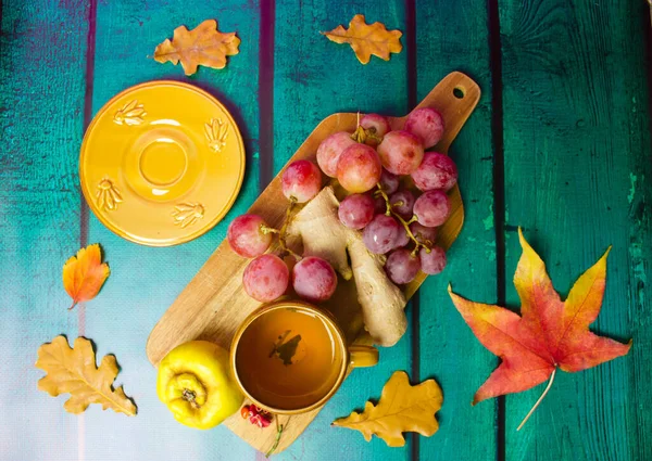 Autumnal still life composition - yellow cup, saucer, grapes, agave, ginger root on cutting board. Fallen oak, maple leaves flatly. Wooden green emerald table background. Bright fall seasonal fruits.