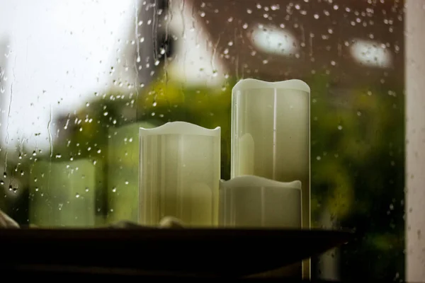 Set of white paraffin artificial tall candles, candlesticks against wet window with water droplets, fall rain outside a window Decor for cozy home. Autumnal cold rainy weather still life. Fall season.
