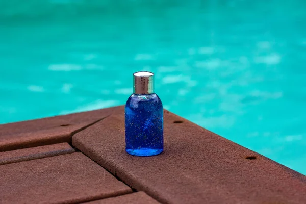 Cosmetic in plastic or glass blue mini bottle in travel version on wooden pier by swimming pool blue water. Natural shower gel, shampoo, liquid soap in jar. Mockup facial skincare cosmetics product.
