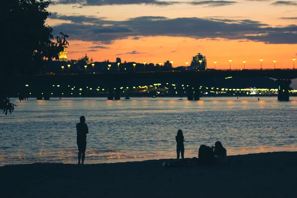 Kyiv at night. The capital of Ukraine plunges into darkness. A silhouette of a nighttime cityscape and sunset orange blue sky. People standing on a shore, Dnipro riverbank looking into a distance.