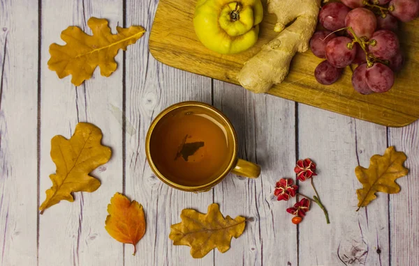 Autumnal still life - a cup of tea on a wooden table among the fallen dry oak leaves flatly. Seasonal fruits - grapes, ginger root, agave. Fall season. Comfort home, atmosphere of warmth and coziness