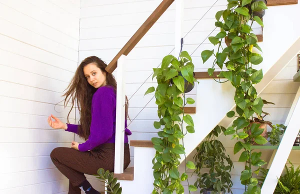 stock image Beautiful stylish long-haired girl with brown hair sits on steps of two-story house, a duplex apartment in a modern white interior. Happy contented young woman in sweet home garden among green plants.