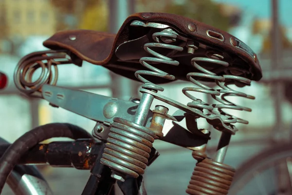 ブラウンレザーヴィンテージ自転車サドル 金属キーキースプリングと古い革自転車クッション 市内の乗り物のためのヴィンテージレトロな交通手段は 都市の通りのぼやけた背景に駐車されています — ストック写真