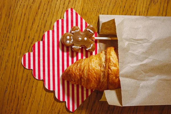 Gingerbread Man and a freshly baked French croissant in a paper bag on a red and white striped napkin on a wooden table top view. A delicious breakfast at home. Delicious Christmas food, treats sweets