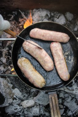 Izgara sosis. İştah açıcı sosisler, sosisler, piknik ateşi manzarasında siyah ızgarada kızartılmış etler. Park ormanlarında dışarıda yemek pişirmek. Kömürde barbekü, açık ateşte sokak ızgarası