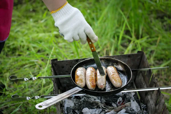 Grilling row sausages. Appetizing sausage, wieners, meats fried on griddle on picnic fire. Cooking outdoors in the park woods. Barbecue on coals, street grill on open fire. A hand in cooking gloves.