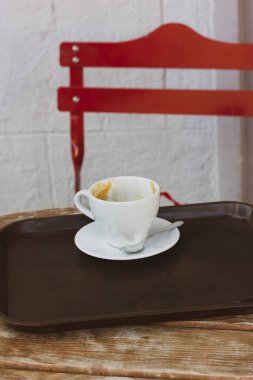 Bir kafenin terasındaki ahşap bir masanın üzerindeki çay tabağının üzerinde beyaz bir kahve fincanı, kentsel bir çevrede bir kahve dükkanı. Sıcak bir içecek ile kavramsal dikey fotoğraf. Kırmızı sandalye.