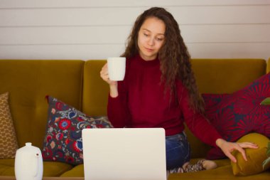 Uzun kıvırcık saçlı genç bir kadın evinin konforunda beyaz bir fincan kahve ile laptopunun önünde çalışıyor ya da dinleniyor. Sonbahar günü evde oturup film izleyen tatlı bir kız..