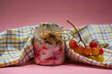 Dry delicious breakfast granola muesli yogurt in glass jar on pink background next to checkered kitchen towel and bunch of grapes. Still life with healthy food, diet, nutrition concept. Dairy product.