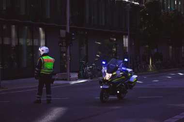 Berlin, Almanya - 1 Ekim 2022: Kasklı bir polis memuru, üzerinde polis resmi olan bir ceket, modern bir motosikletin yanındaki karanlık şehir caddesinde devriye geziyor. Şehirlerdeki kolluk kuvvetleri. Suç vakası