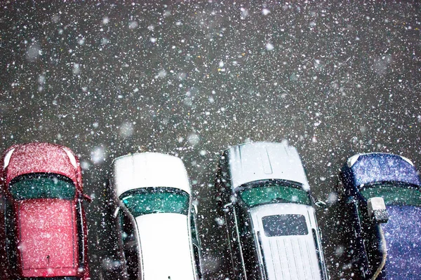 Cars parked in a private parking lot in the yard of neighborhood in the snowfall. Roofs of passenger cars view from above. Snowflakes falling down. Frosty cold snowy day. First snow in December.