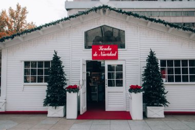 Madrid, İspanya. 30 Kasım 2022 Noel Baba 'nın beyaz ahşap evi. Yeni yıl için ev dekorasyonu ürünleri ve sokakta Noel ağaçları. Tienda de Navidad yazılı kırmızı masa