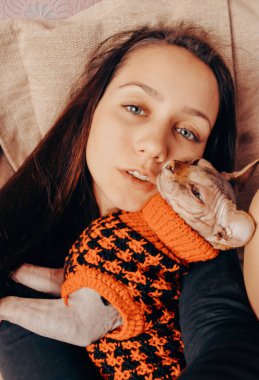 Genç bayan, kız evinde kırmızı kazaklı Kanadalı bir Sphynx kedisine sarılıyor. Sevilen bir evcil hayvanla temas, etkileşim. Sevgili tüysüz kedicik. Hayvanlarla aşk paylaşımı. Sfenks modası