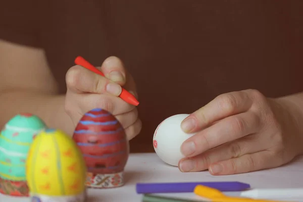Coloring Easter eggs with red wax crayon in home kitchen. White Easter egg in women's hands before dyeing colors. Easter egg festival. Artistic work, handmade DIY. People preparing for Easter in April