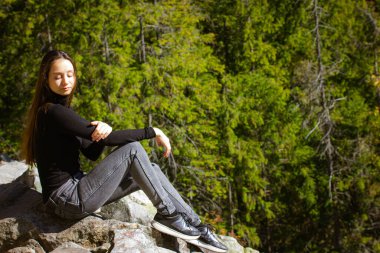 Yeşil kozalaklı ormanda dağın tepesinde oturan uzun saçlı zarif genç bir kadın. Ormanda yürüyüş yapan hassas bir kız. Macera, yolculuk. Güneşli bir sonbahar gününde doğada gezmenin tadını çıkarıyorum..