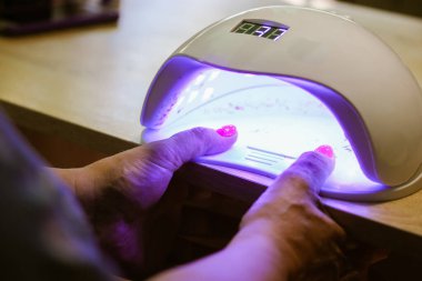 Kadın elleri mor ötesi lambanın içinde jel metoduyla pembe tırnakları kurutmak için, plaka kaplaması için. Manikür işlemi. Güzellik salonunda yeni uygulanan jel ojeleri ya da hibrit ojeler, güzellik uzmanı. 