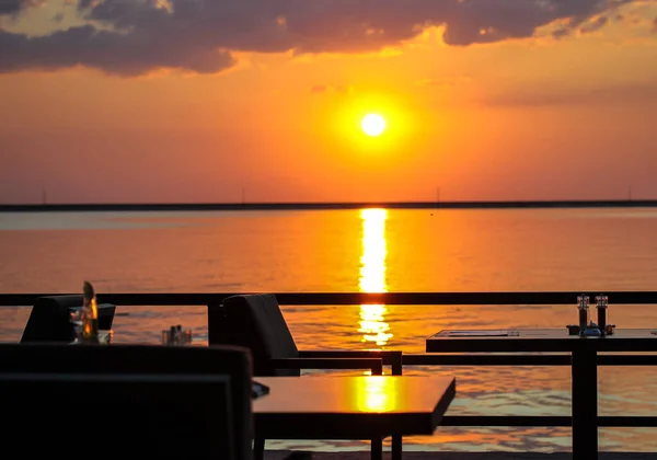 Silhouettes of tables and chairs on cafe restaurant terrace against background of red yellow-orange sky at sunset. The sun setting over a horizon. Beautiful tranquil seascape. Vacation trip concept.