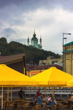 Kyiv, Ukraine. July 1, 2021. Exterior of McDonald's fast food cafe. Yellow umbrellas outside, people eating sitting at a table. City architecture against cloudy sky before rain. St. Andrew's Church