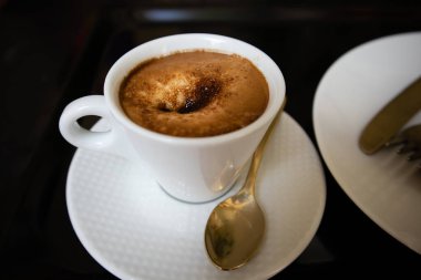 Porcelain white coffee cup with cappuccino, mochaccino on a saucer with a teaspoon on a black marble table to view. Hot tasty drink for a breakfast, branch, lunch. Aromatic beverage on a kitchen.