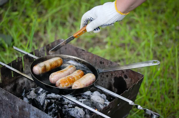 Grilling row sausages. Appetizing sausage, wieners, meats fried on griddle on picnic fire. Cooking outdoors in the park woods. Barbecue on coals, street grill on open fire. A hand in cooking gloves.