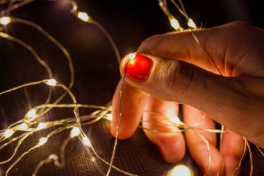 Karışık bir çelenk tutan kadın el, parlayan ışıklar. 2023 yılbaşı arifesi için bir Noel ağacı süslemeye hazırlanan kadın. Turuncu-kırmızı manikürlü ince parmaklar koyu arkaplanda bir tel tutuyor.