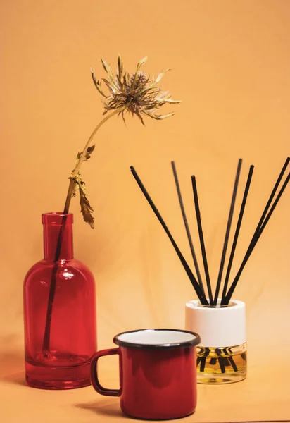 Stylish modern still life with red glass vase, dried flower, red coffee cup mug, aroma diffuser oil with black bamboo sticks on brown and beige vertical background. Cozy home interior decor.
