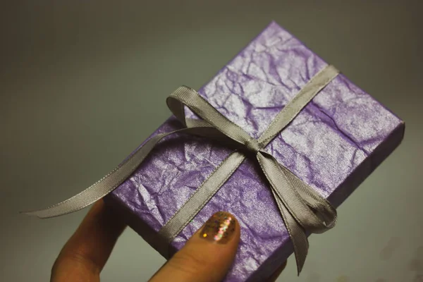 Purple gift box with silver silk bow in woman\'s hand on gray background. Gift giving. Shopping, shopping, gifts to loved ones concept. Gifts in a box for the lovers, mother, daughter, girl Boxing gift