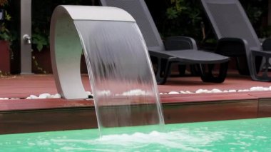 Stainless steel swimming pool wall waterfall jet on luxury resort. Water feature fountain with clean green water falling down under high pressure. Lounge area with sun loungers by a pool in summer.