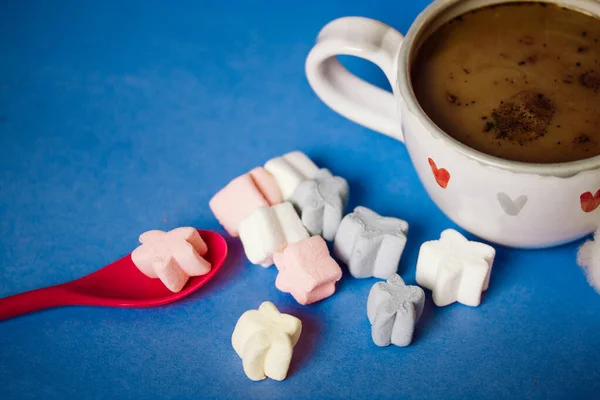Small cup of coffee or cocoa with hearts on a blue background view from above flatly. A pink spoon and lots of marshmallows scattered on the table. Sweet table, dessert. Cozy still life with hot drink