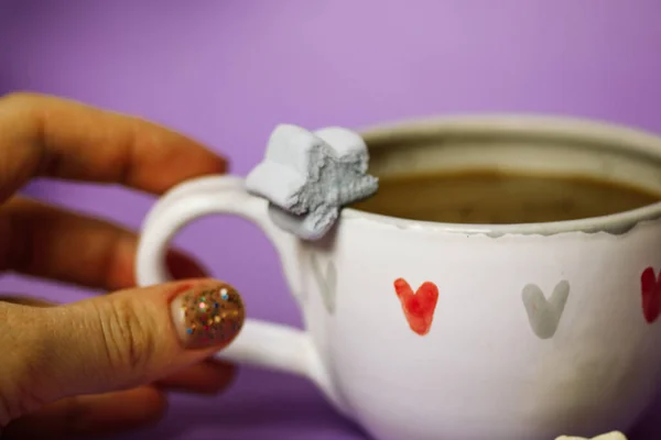 A cup of coffee, cocoa, chocolate with hearts on a violet background in female hand. Marshmallows on a table. Cozy still life with hot drink made with love and marshmallow. Happy Valentines Day.
