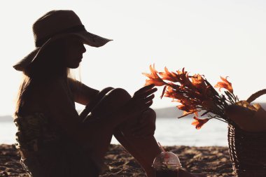 Silhouette of teenage girl in straw hat portrait in profile touching garden orange lily flowers with a hand. Female child on vacation in nature, picking flowers on a sandy beach coast on summer sunset