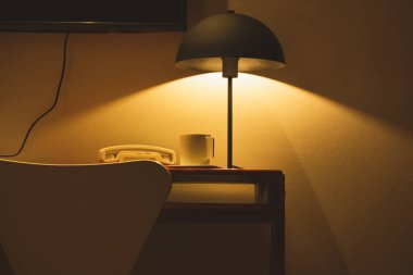 A vintage retro stylish table lamp, cable phone, white coffee or tea cup stand on a table under a muted warm yellow light. A cozy, homely atmosphere in the evening dark darkness Modern bedroom indoors