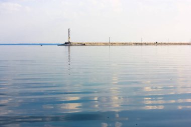 Lighthouse glowing in a distance. A concrete breakwater on a horizon. A beautiful seascape. A blue sky and reflection in the water water without waves at daytime. Anti-wave protection. Calmness relax.
