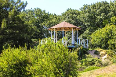 Beyaz ahşap çardak, pavyon ya da turuncu tavanlı orkestra sehpası yeşil güzel bir halk parkında, yazın veya ilkbaharda güneşli bir günde şehir bahçesinde. Çalıların, ağaçların üzerindeki yemyeşil yapraklar. Rahatlamak için huzurlu bir yer..